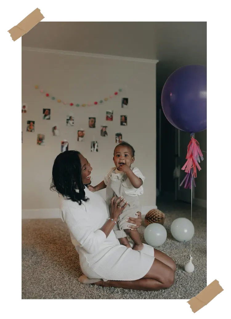 A joyful mother kneeling on the floor, holding her baby during a first birthday celebration, surrounded by balloons and family photos on the wall.