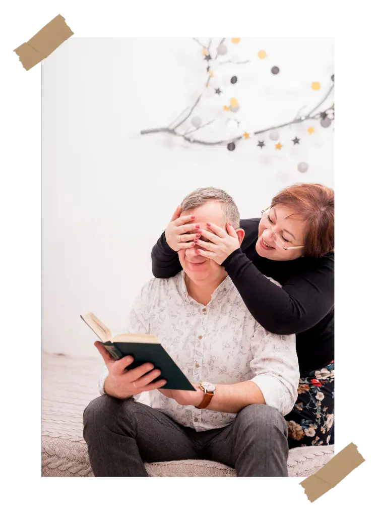 A woman playfully covering a man's eyes as he reads a book, symbolizing cherished memories and the joy of storytelling.